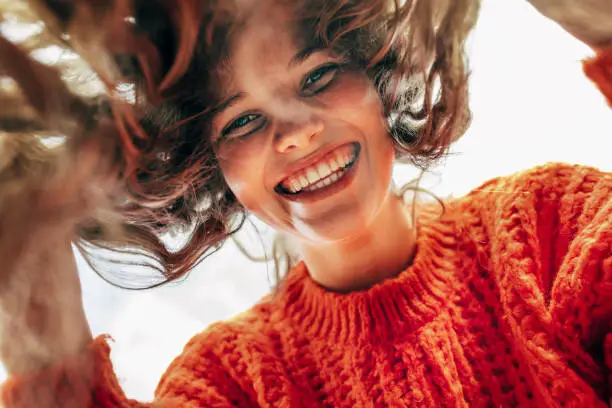 Bottom view of a candid beautiful young woman wearing a knitted orange sweater smiling broadly and looking directly to the camera outdoors. The pretty female has joyful expression, resting in the park