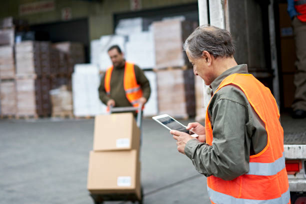 Senior Male Transportation Foreman Using Digital Tablet Focus on foreground man in casual clothing and reflective vest checking portable information device as coworker moves boxes with hand truck in background. loading bay stock pictures, royalty-free photos & images