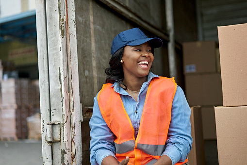 Waist-up front view of mid 20s transportation worker sitting on back of loaded truck and laughing as she takes a break with off-camera loading dock worker.