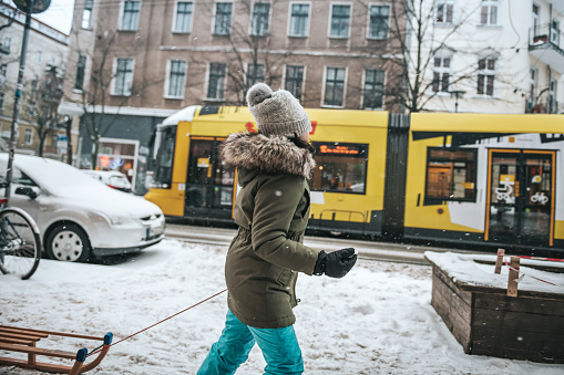 little girl pulling sleigh in snowy streets of central berlin while yellow tram is passing by