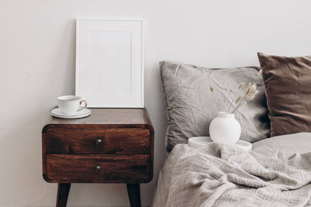 istock Vertical white picture frame mockup. Retro wooden bedside table. Modern white ceramic vase with dry Lagurus ovatus grass and cup of coffee. Beige linen, velvet pillows in Scandinavian bedroom. 1338626077