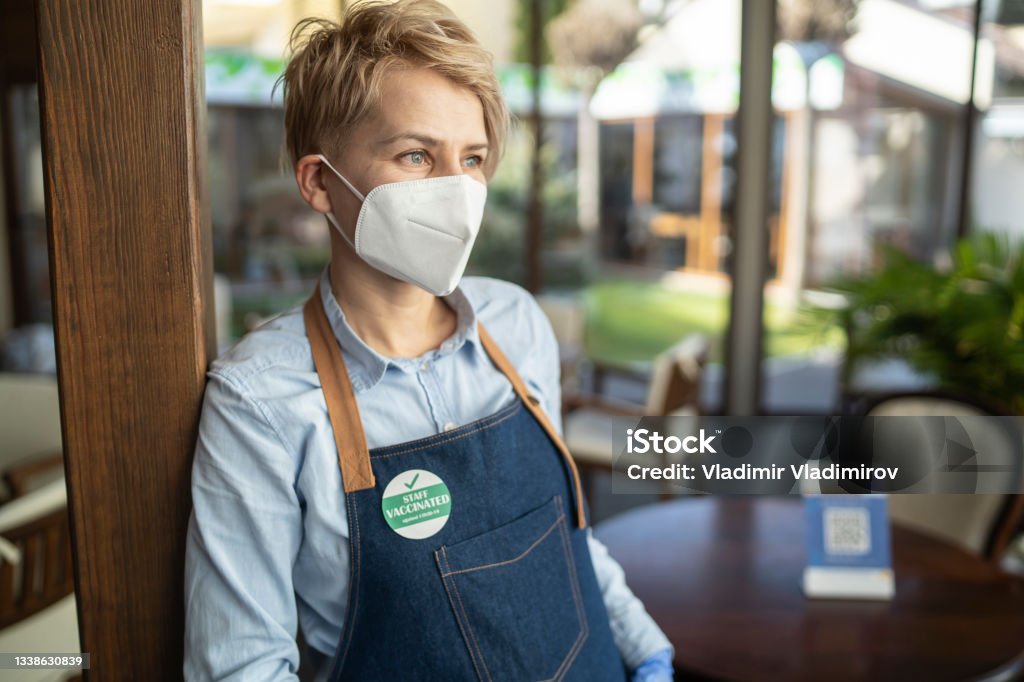 Portrait of a female restaurant owner wearing a face mask and standing in front of her restaurant Female restaurant owner  with face protection and badge showing vaccination against COVDI-19 standing in front of her restaurant and smiling. Waitress Stock Photo