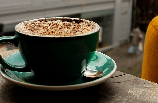 Stock photo showing a close-up view of frothy, hot chocolate in a green cup and saucer with a metal teaspoon. The hot drink is located on a wooden rustic table beside a window overlooking a street scene.