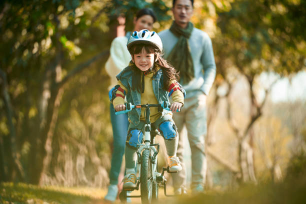 little asian girl riding bike in park with parent watching from behind stock photo