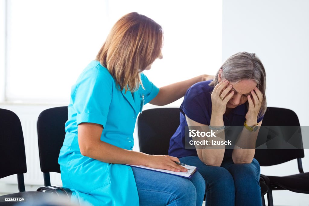 Depressed senior woman talking with nurse Nurse consoling an elderly lady. They are sitting on chairs in waiting room in the hospital. Hospital Stock Photo