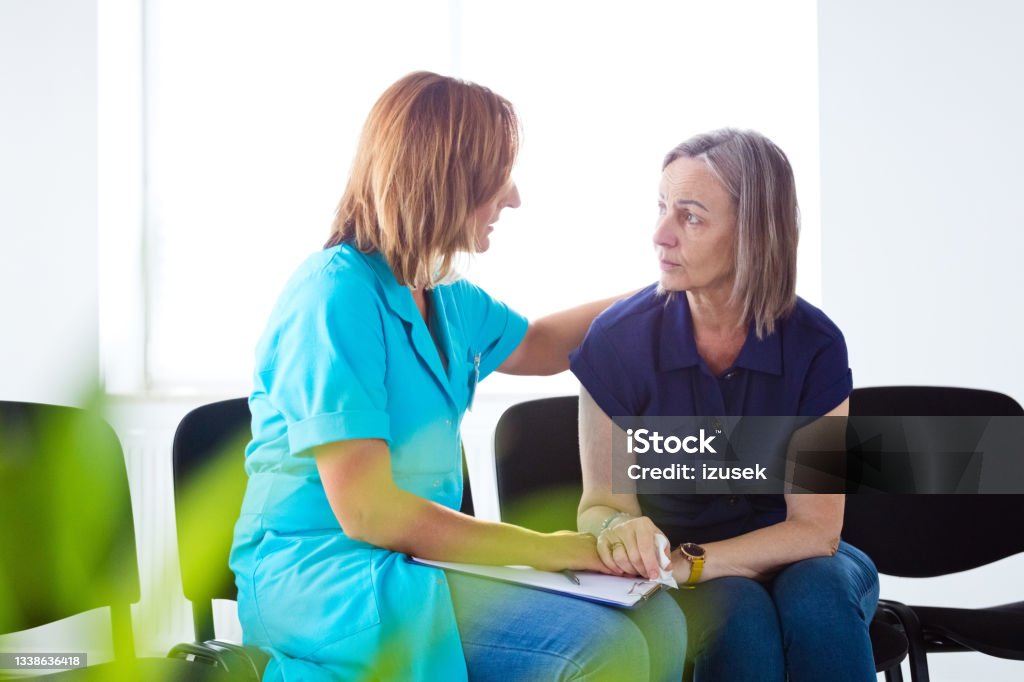 Worried senior woman talking with nurse Nurse consoling an elderly lady. They are sitting on chairs in waiting room in the hospital. Consoling Stock Photo