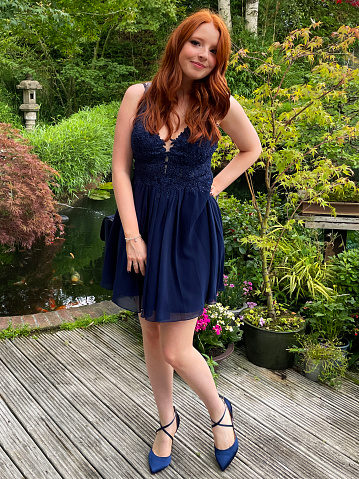 Stock photo showing a beautiful, ginger haired young woman looking at camera relaxed and smiling modelling a dark blue, short cocktail prom dress on garden decking besides a pond.