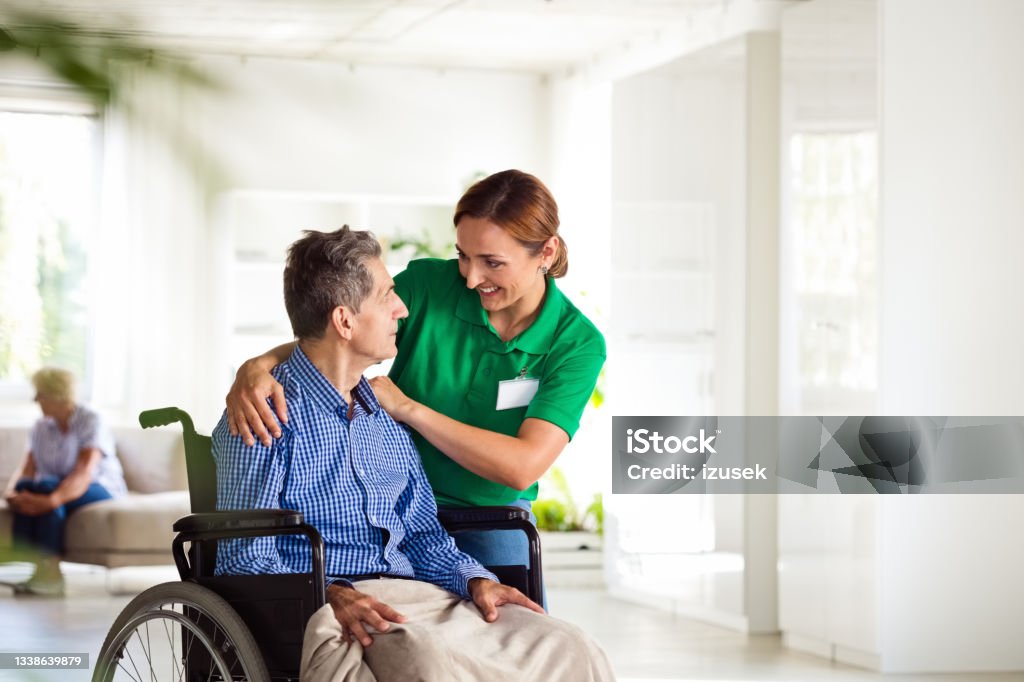 Cheerful social worker supporting senior man Elderly man in retirement home. Friendly nurse talking with worried senior man sitting in wheelchair. 70-79 Years Stock Photo