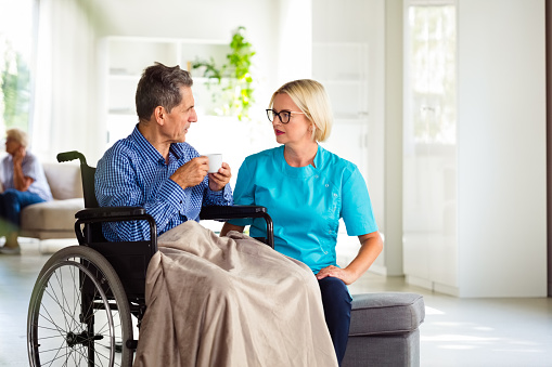 Friendly home nurse talking senior man sitting in wheelchair. They are in nursing home.