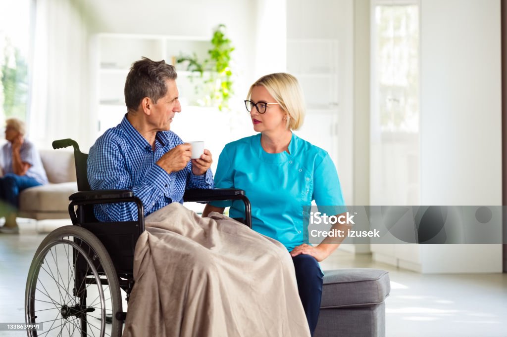 Nurse talking with senior man in retirement house Friendly home nurse talking senior man sitting in wheelchair. They are in nursing home. Assistance Stock Photo