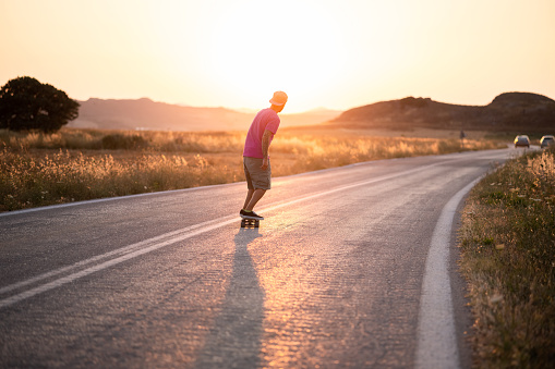 Young Caucasian man wearing a pink t-shirt enjoying himself outdoors, riding his skateboard on an empty open road