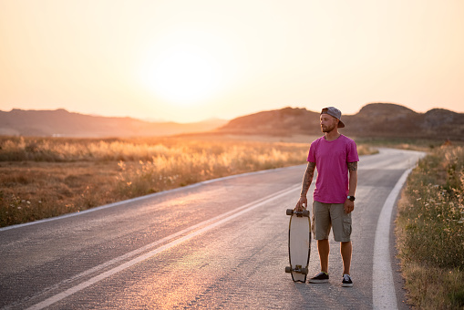 Young Caucasian man wearing a pink t-shirt enjoying himself outdoors, holding his skateboard on an empty open road