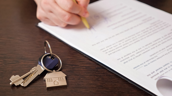 Man client signs new apartment purchase contract with pen near keys with house shaped breloque put on table extreme close view