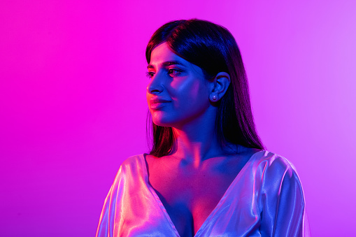 Tenderly looking away. Cropped portrait of young beautiful woman isolated over gradient pink blue purple background in neon lights. Youth culture. Concept of human emotions, facial expression, ad