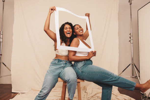 Best friends laughing through a white picture frame Best friends laughing through a white picture frame. Two happy female friends having fun together while wearing denim jeans and bras. Two self-confident young women being their natural selves. body confidence stock pictures, royalty-free photos & images