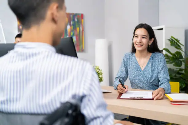 Photo of A job interview with a disabled man in wheelchair, beautiful woman of Asian Korean beauty asks questions to boy in shirt, she notes the answers, they are sitting in company office on two sides of desk