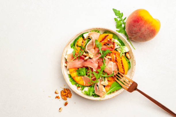 istock Salad bowl with grilled peach, prosciutto, goat cheese, walnuts and arugula on white background, top view, copy space 1338662699
