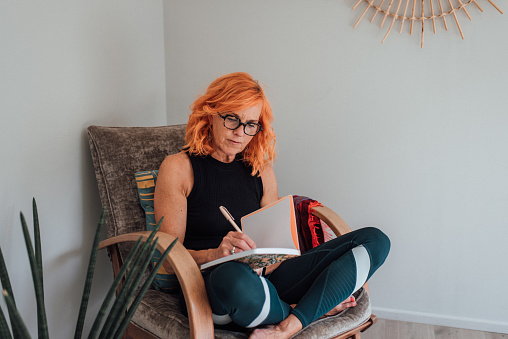 Orange haired pilates instructor sitting at home writing notes in her journal preparation for her online class