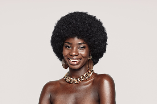 Portrait of beautiful young African woman in golden jewelry looking at camera and smiling while standing against gray background