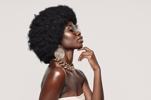 Side view of beautiful young African woman in golden jewelry keeping eyes closed and touching face while standing against gray background