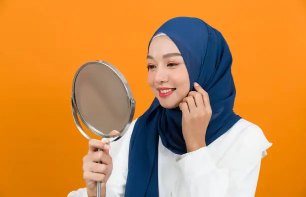 Asian young Muslim woman wearing hijab headscarf smiling and looking at mirror on orange colour background in studio