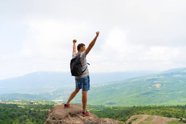 istock Young man standing on top of hill with hands up wearing gray backpack. Young guy raising hands up in nature.  Concept of travel lifestyle and vacation in mountains 1338687488