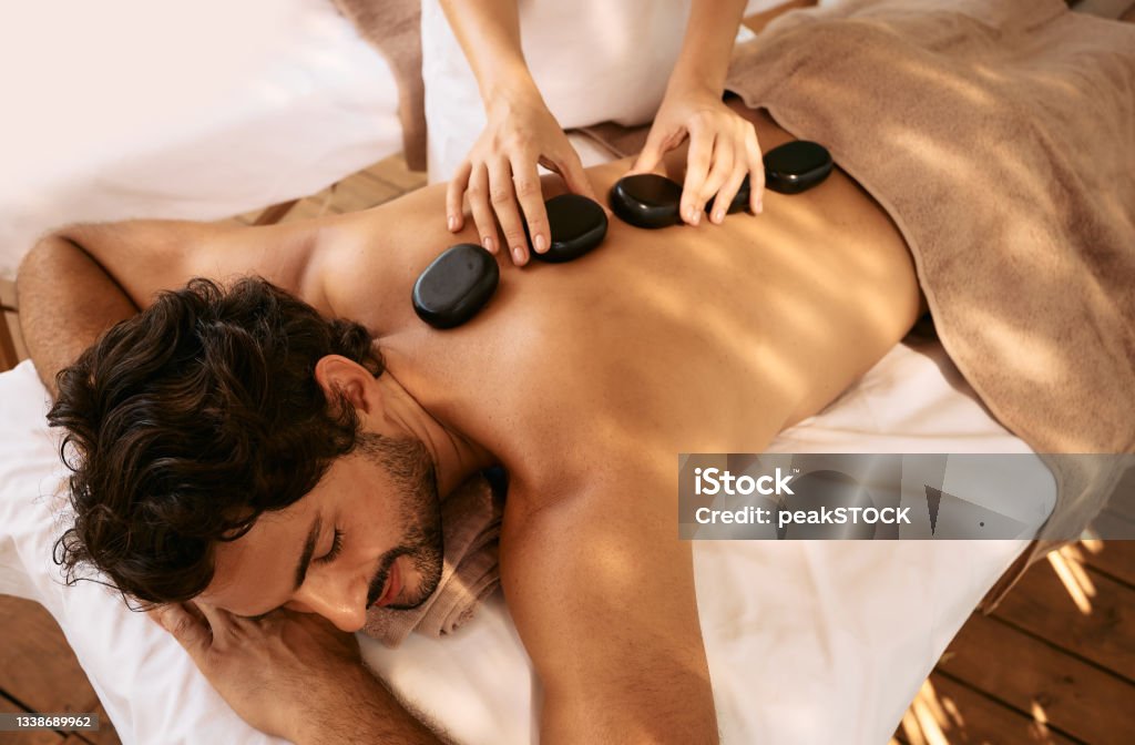 Handsome man at spa resort receives hot stone massage. Hot stone massage therapy using smooth, flat, heated stones Massaging Stock Photo