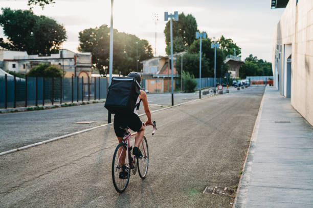 Giovane donna di consegna adulta sta andando in bicicletta per consegnare del cibo al tramonto - foto stock