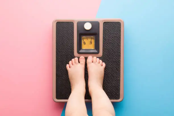 Photo of Baby barefoot standing on weight scales on light pink blue floor background. Pastel color. Closeup. Care about body. Children weight control concept. Top down view. Two sides.