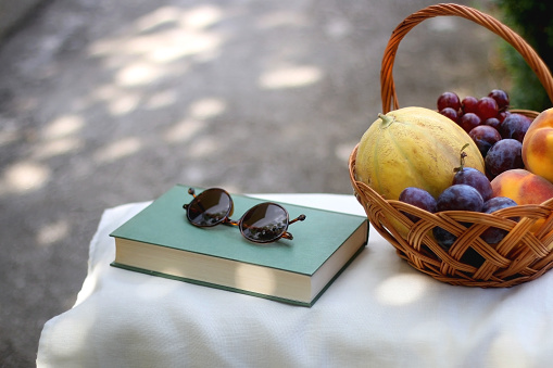 Vintage basket full of various fruit, hardback book and retro sunglasses on picnic basket. Selective focus.