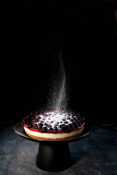 Image of icing sugar being sifted onto blackcurrant cheesecake with buttery biscuit base on black cake stand, black background, white powder falling and floating mid-air, focus on foreground, copy space