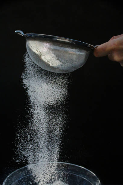 Image of unrecognisable person's hand holding a metal sieve, white flour being sifted against a black background, white powder falling and floating mid-air, focus on foreground stock photo