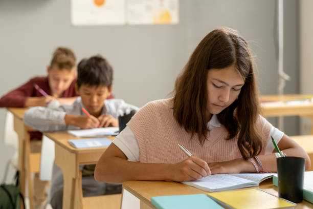 istock Cute schoolgirl making notes in copybook while bending over desk 1338720138