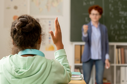 Back view of African schoolgirl raising hand at lesson while teacher by blackboard pointing at her