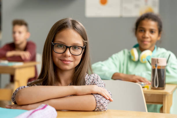 Cute schoolgirl in eyeglasses looking at you by desk stock photo