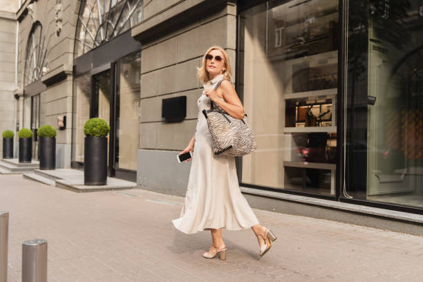 beautiful blonde caucasian mature woman walking while shopping on the city street, carrying handbag outdoors. middle-aged businesswoman buying goods clothes - purse bag glamour personal accessory imagens e fotografias de stock