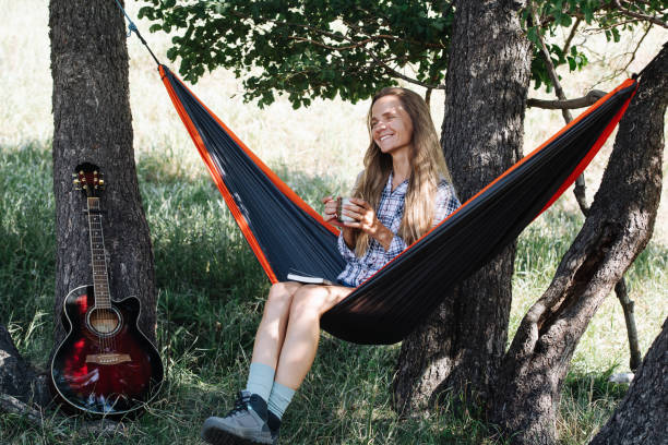 istock Joyful woman sitting in a hammock, between two big trees. Drinking tea 1338725527
