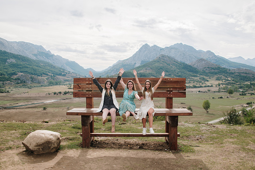 Three female friends with arms raised sitting on a giant bench in a mountains area