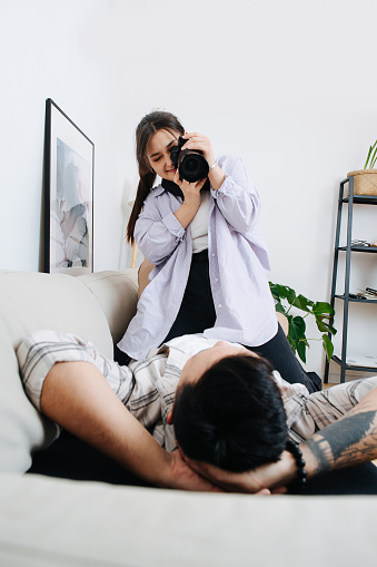 Intimate couple, man lying on a sofa, relaxing in a living room, woman sitting on him, taking photo on a digital camera.