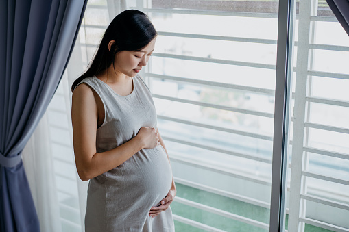 Image of a smiling Asian Chinese pregnant woman standing and touching her belly at home