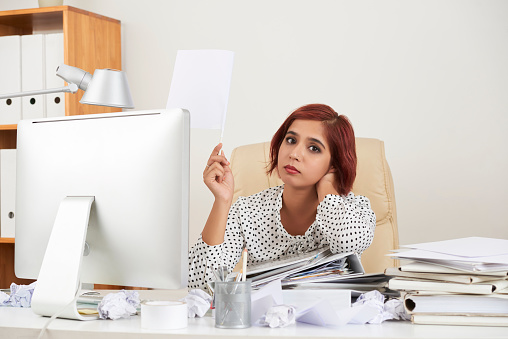 Tired hopeless pretty young businesswoman leaning on stack of documents and lifting hand with white flag