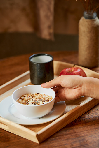 Close-up image of woman making breakfast and putting bowl of granola, apple and cup of coffee on tray
