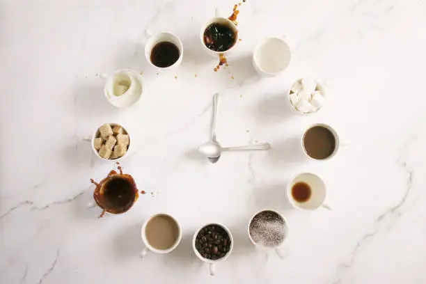 Photo of Image of white, espresso cup clock face design , sugar cubes, milk, coffee and coffee beans in cups, teaspoon clock hands, overflowing, spraying, splashing droplets floating mid-air,  marble effect background, elevated view, copy space