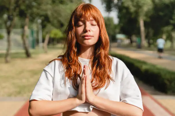 Photo of Young redhead caucasian woman student teenager girl meditating warming-up before training, feeling zen-like while jogging running outdoors in park. Active lifestyle concept.