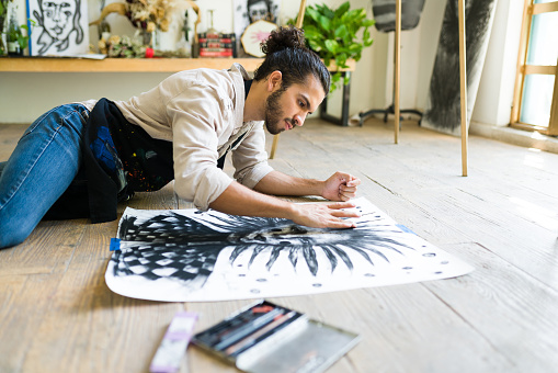 Attractive hispanic painter drawing an artistic charcoal portrait while lying on the art studio floor