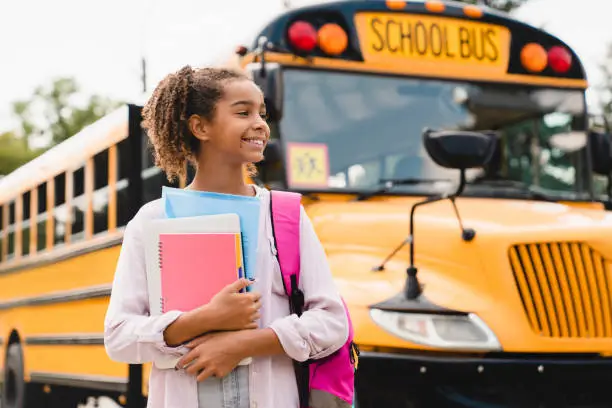 African-american girl teenager pupil student preparing to go to school after summer holidays holding books and notebooks standing next to the school bus.
