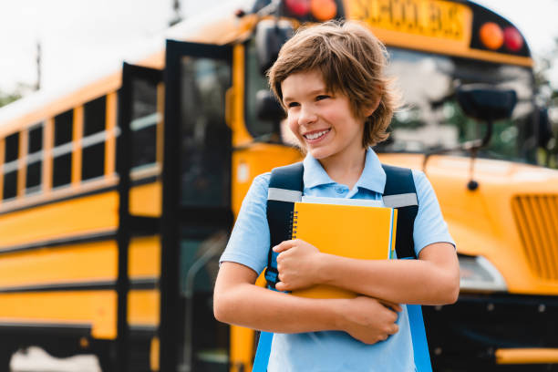 Young caucasian schoolboy kid pupil student holding copybooks and books preparing for school standing next to the school bus. Back to school concept Young caucasian schoolboy kid pupil student holding copybooks and books preparing for school standing next to the school bus. Back to school concept first grade classroom stock pictures, royalty-free photos & images