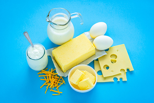 High angle view of a composition of main dairy products shot on blue background that includes butter, milk, yogurt, cheese and eggs. The composition is at the center of an horizontal frame. High resolution 42Mp studio digital capture taken with Sony A7rII and Sony FE 90mm f2.8 macro G OSS lens