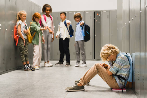 istock Sad crying schoolboy pupil sitting on the floor at the school hall while his classmates teenagers laughing at him, bullying and torturing him. Social exclusion problem 1338738807