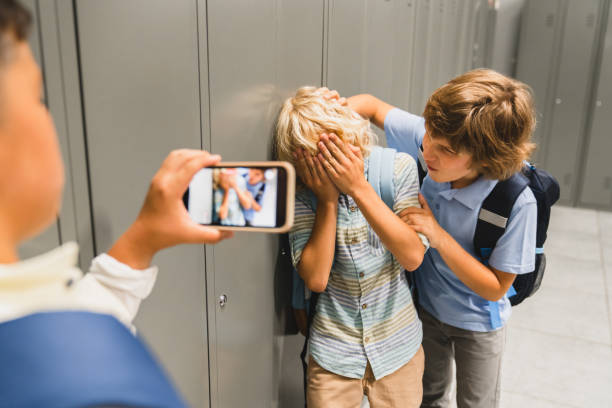 schulkinder, die grausame jungen am telefon filmen, wie sie ihren klassenkameraden in der schulhalle foltern. pubertät schwieriges alter - vorurteil stock-fotos und bilder